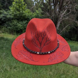 May include: A red felt fedora hat with a brown beaded band and a tree and leaf design.
