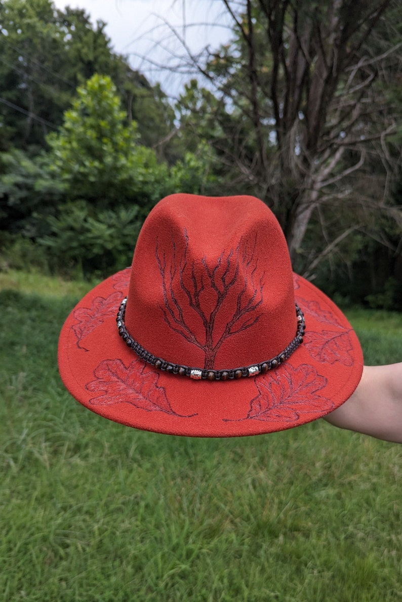 May include: A red felt fedora hat with a brown beaded band and a tree and leaf design.