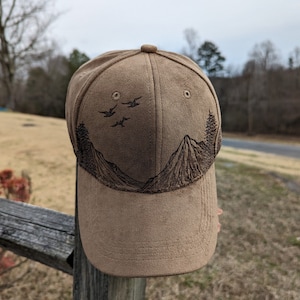 May include: A brown suede baseball cap with an embroidered design of mountains and birds. The cap has a curved brim and a brown adjustable strap.