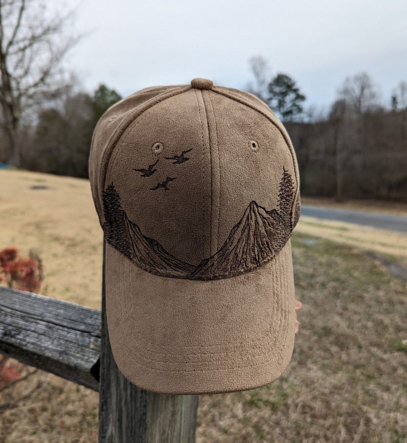 May include: A brown suede baseball cap with an embroidered design of mountains and birds. The cap has a curved brim and a brown adjustable strap.