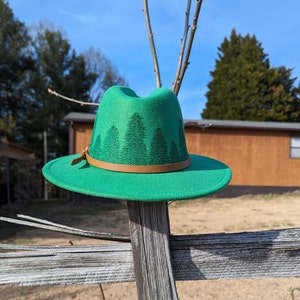 May include: A green felt fedora hat with a brown leather band and a forest silhouette design. The hat is sitting on a wooden fence.