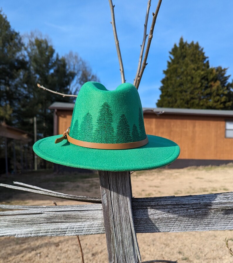 May include: A green felt fedora hat with a brown leather band and a forest silhouette design. The hat is sitting on a wooden fence.
