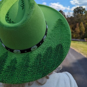Puede incluir: Un sombrero fedora verde brillante con una banda negra con adornos plateados. El ala del sombrero est&aacute; decorada con una escena de bosque en tonos verdes. El sombrero tiene un dise&ntilde;o de hojas en el lateral.
