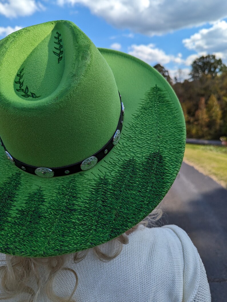 Puede incluir: Un sombrero fedora verde brillante con una banda negra con adornos plateados. El ala del sombrero est&aacute; decorada con una escena de bosque en tonos verdes. El sombrero tiene un dise&ntilde;o de hojas en el lateral.