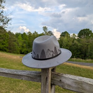 Op de afbeelding: Een grijze wollen fedora hoed met een zwarte band en een bergketen ontwerp. De hoed ligt op een houten hek.