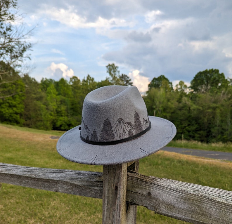 Op de afbeelding: Een grijze wollen fedora hoed met een zwarte band en een bergketen ontwerp. De hoed ligt op een houten hek.