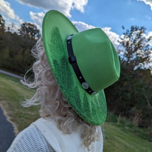 Puede incluir: Un sombrero fedora verde brillante con una banda negra y tachuelas plateadas. El ala presenta un dise&ntilde;o texturizado en verde oscuro. El sombrero se usa al aire libre, con un cielo azul y &aacute;rboles de fondo.