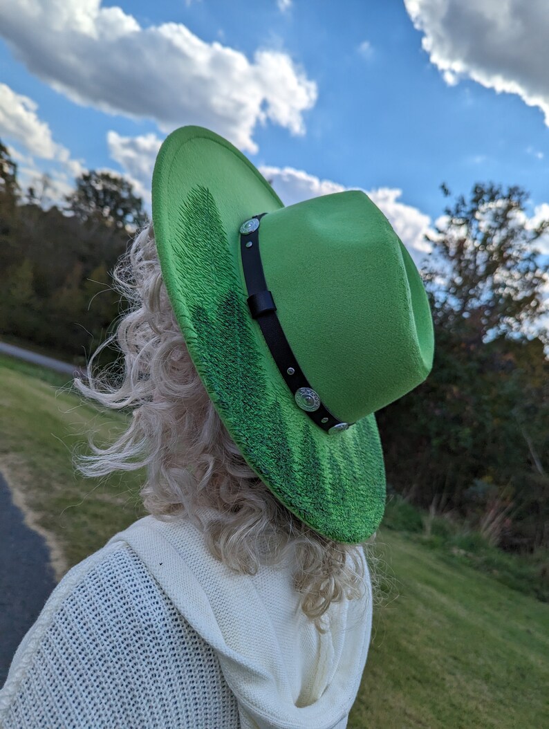 Puede incluir: Un sombrero fedora verde brillante con una banda negra y tachuelas plateadas. El ala presenta un dise&ntilde;o texturizado en verde oscuro. El sombrero se usa al aire libre, con un cielo azul y &aacute;rboles de fondo.