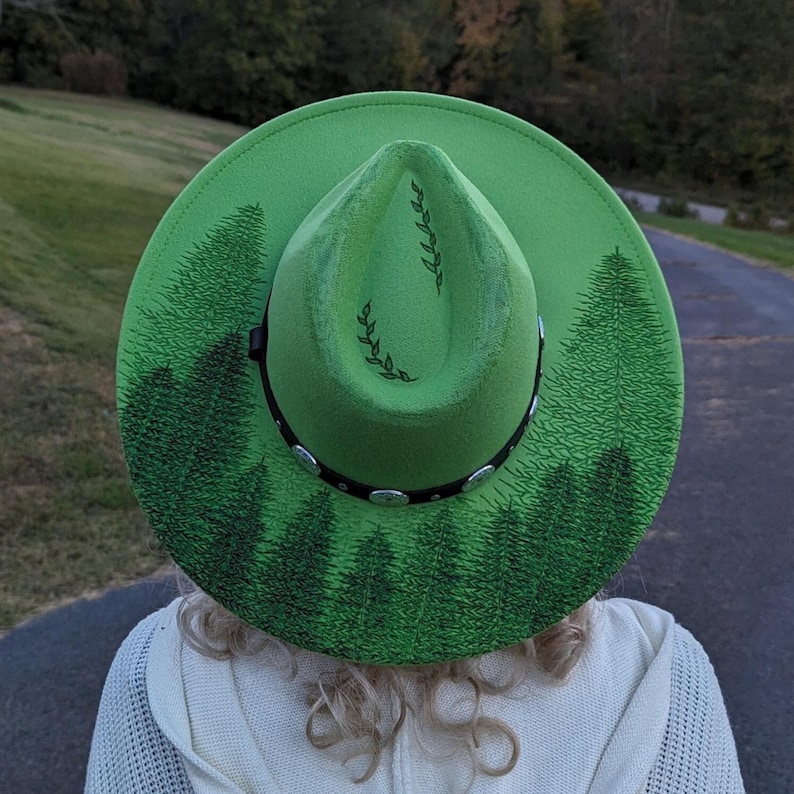 Puede incluir: Un sombrero fedora verde brillante con una banda negra y tachuelas plateadas. El sombrero presenta un dise&ntilde;o de bosque pintado en verde oscuro alrededor del ala y en la corona. Un accesorio elegante.