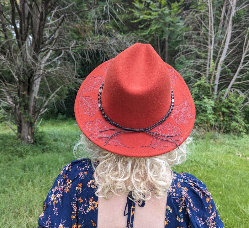May include: A red felt hat with a wide brim and a brown leather band. The brim is decorated with a white leaf design.
