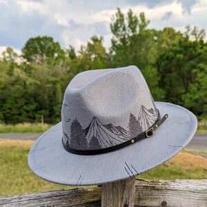Op de afbeelding: Een grijze wollen fedora hoed met een zwarte leren band en een berglandschap ontwerp. De hoed heeft een bruine leren gesp en gouden klinknagels.