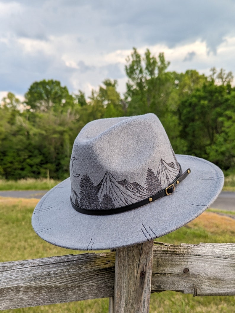 Op de afbeelding: Een grijze wollen fedora hoed met een zwarte leren band en een berglandschap ontwerp. De hoed heeft een bruine leren gesp en gouden klinknagels.