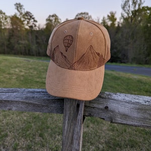 K&ouml;nnte beinhalten: Eine braune Baseballkappe aus Wildleder mit einem gestickten Design von Bergen und einem Hei&szlig;luftballon.