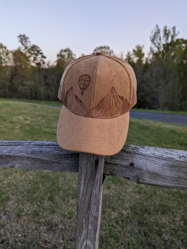 K&ouml;nnte beinhalten: Eine braune Baseballkappe aus Wildleder mit einem gestickten Design von Bergen und einem Hei&szlig;luftballon.
