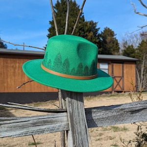 May include: A green fedora hat with a brown band and a forest print design. The hat is sitting on a wooden fence.