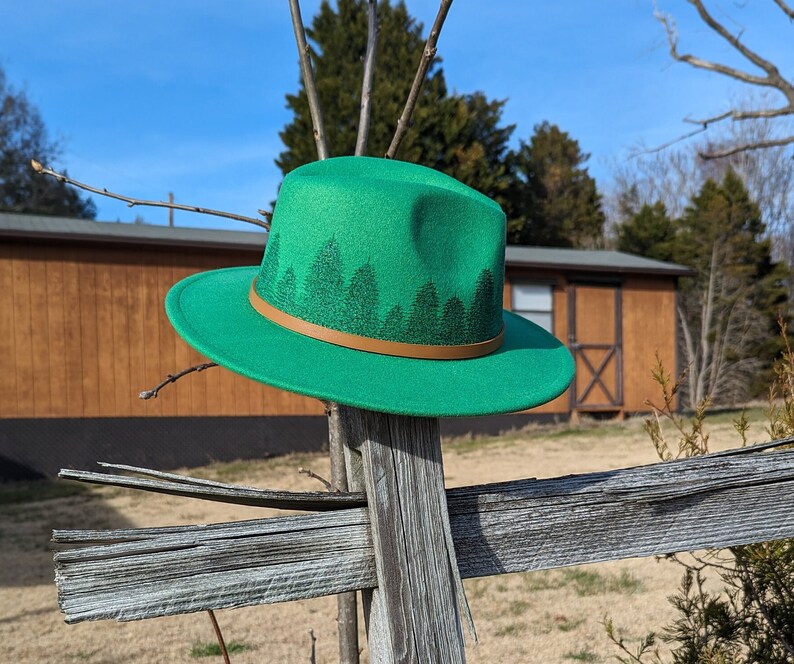 May include: A green fedora hat with a brown band and a forest print design. The hat is sitting on a wooden fence.