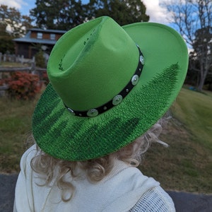 Puede incluir: Un sombrero de vaquero verde brillante con una banda negra adornada con tachuelas plateadas. El ala presenta un dise&ntilde;o detallado de bosque en varios tonos de verde. Un accesorio elegante.