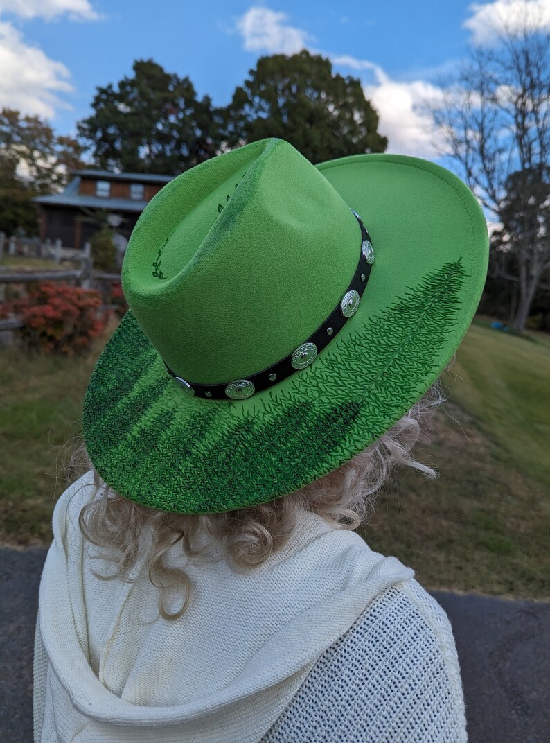 Puede incluir: Un sombrero de vaquero verde brillante con una banda negra adornada con tachuelas plateadas. El ala presenta un dise&ntilde;o detallado de bosque en varios tonos de verde. Un accesorio elegante.