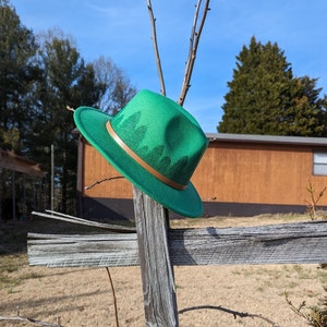 May include: A green fedora hat with a brown leather band and a forest print. The hat is hanging on a wooden fence post.