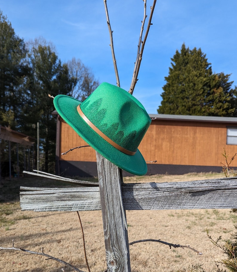 May include: A green fedora hat with a brown leather band and a forest print. The hat is hanging on a wooden fence post.
