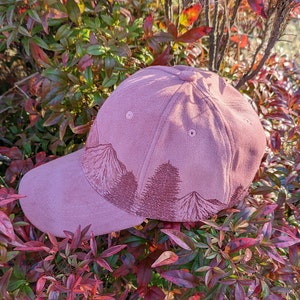 May include: A pink suede baseball cap with an embroidered mountain scene. The cap has a curved brim and a classic six-panel design.