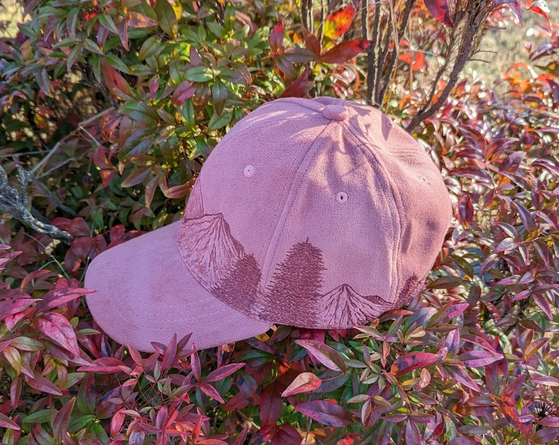 May include: A pink suede baseball cap with an embroidered mountain scene. The cap has a curved brim and a classic six-panel design.