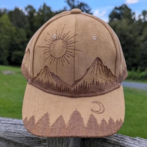 May include: Tan baseball cap with a mountain and sun design. The cap features a detailed illustration of a sun, mountains, and trees in shades of brown. The brim has a repeating tree line pattern, and a crescent moon is on the side.