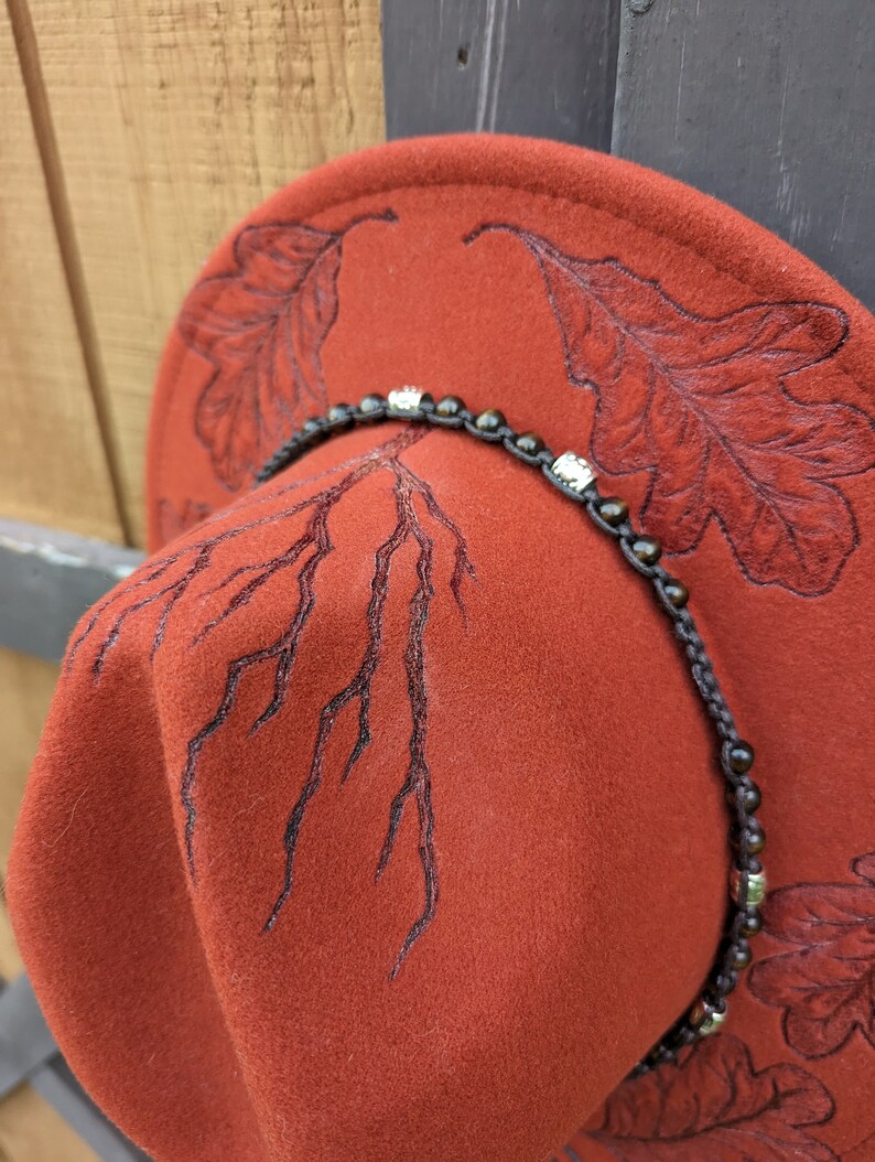 May include: A rust-colored felt hat with a black embroidered design of oak leaves and branches. The hat has a brown leather band with silver and black beads.