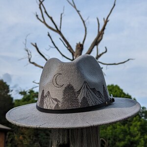 Op de afbeelding: Een grijze wollen fedora hoed met een zwarte leren band en een geborduurd ontwerp van een berg en een halve maan op de kroon.