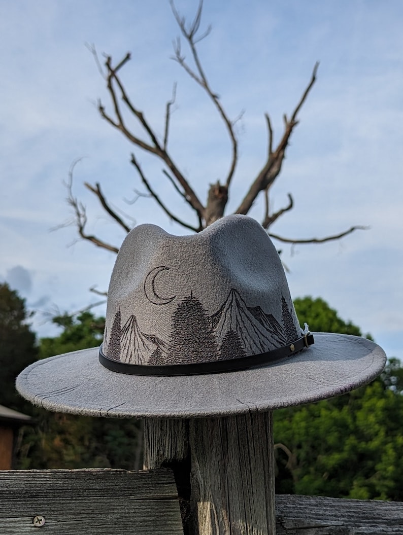 Op de afbeelding: Een grijze wollen fedora hoed met een zwarte leren band en een geborduurd ontwerp van een berg en een halve maan op de kroon.