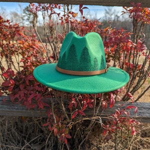 May include: A green felt fedora hat with a brown leather band and a forest silhouette design. The hat is sitting on a wooden fence with red leaves.