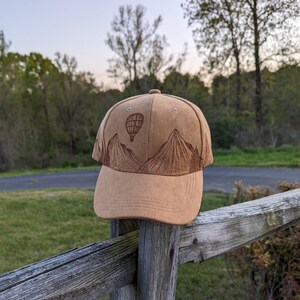 K&ouml;nnte beinhalten: Eine braune Baseballkappe aus Wildleder mit einem gestickten Design von Bergen und einem Hei&szlig;luftballon.