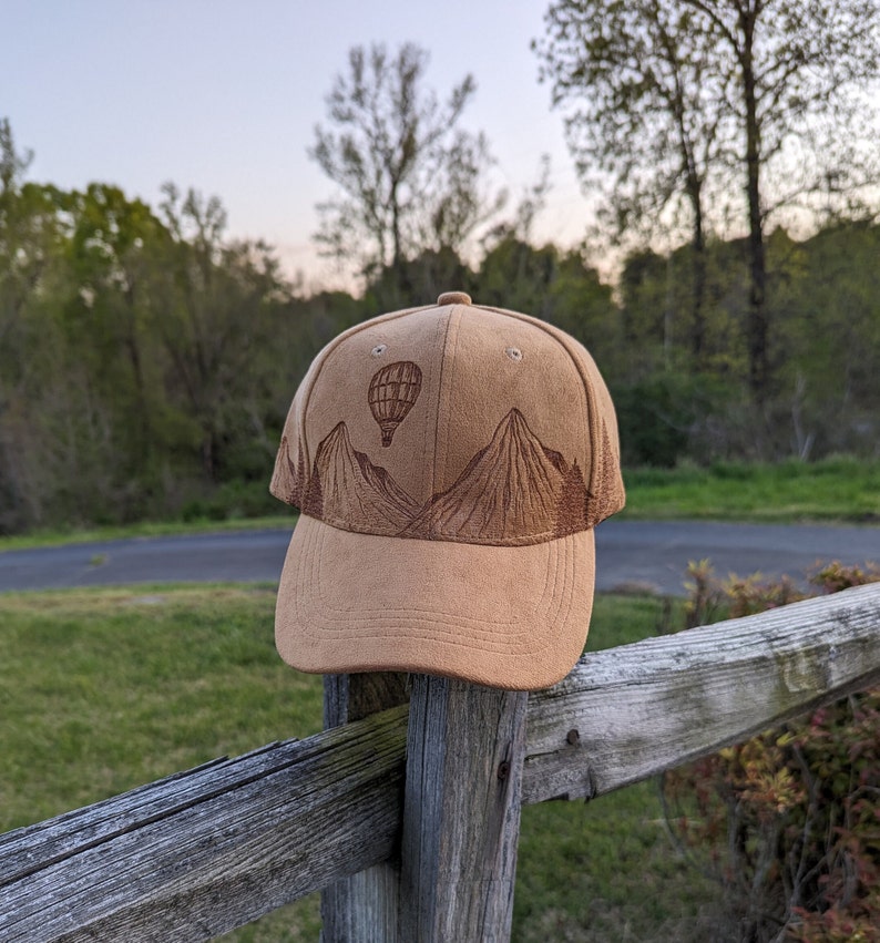 K&ouml;nnte beinhalten: Eine braune Baseballkappe aus Wildleder mit einem gestickten Design von Bergen und einem Hei&szlig;luftballon.