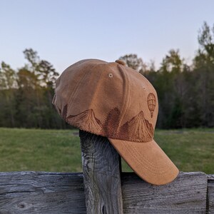 K&ouml;nnte beinhalten: Eine braune Baseballkappe aus Wildleder mit einem gestickten Design aus Bergen und einem Hei&szlig;luftballon. Die Kappe hat einen gebogenen Schirm und ein braunes Band.