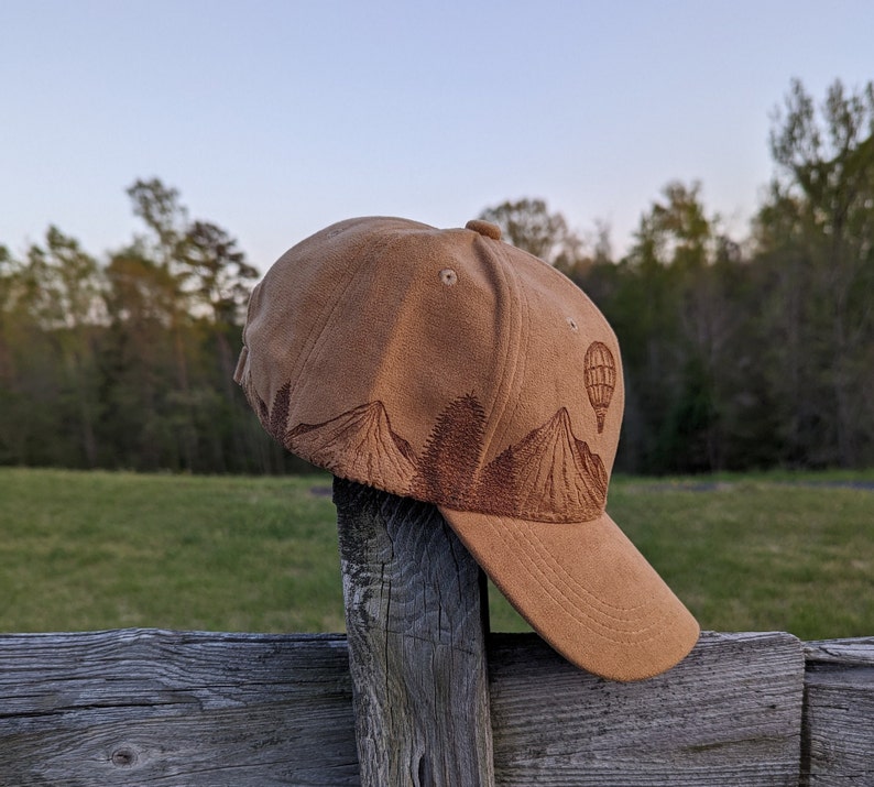 K&ouml;nnte beinhalten: Eine braune Baseballkappe aus Wildleder mit einem gestickten Design aus Bergen und einem Hei&szlig;luftballon. Die Kappe hat einen gebogenen Schirm und ein braunes Band.