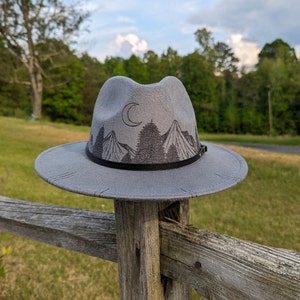 Op de afbeelding: Een grijze wollen fedora hoed met een zwarte band en een geborduurd ontwerp van bergen, bomen en een halve maan.