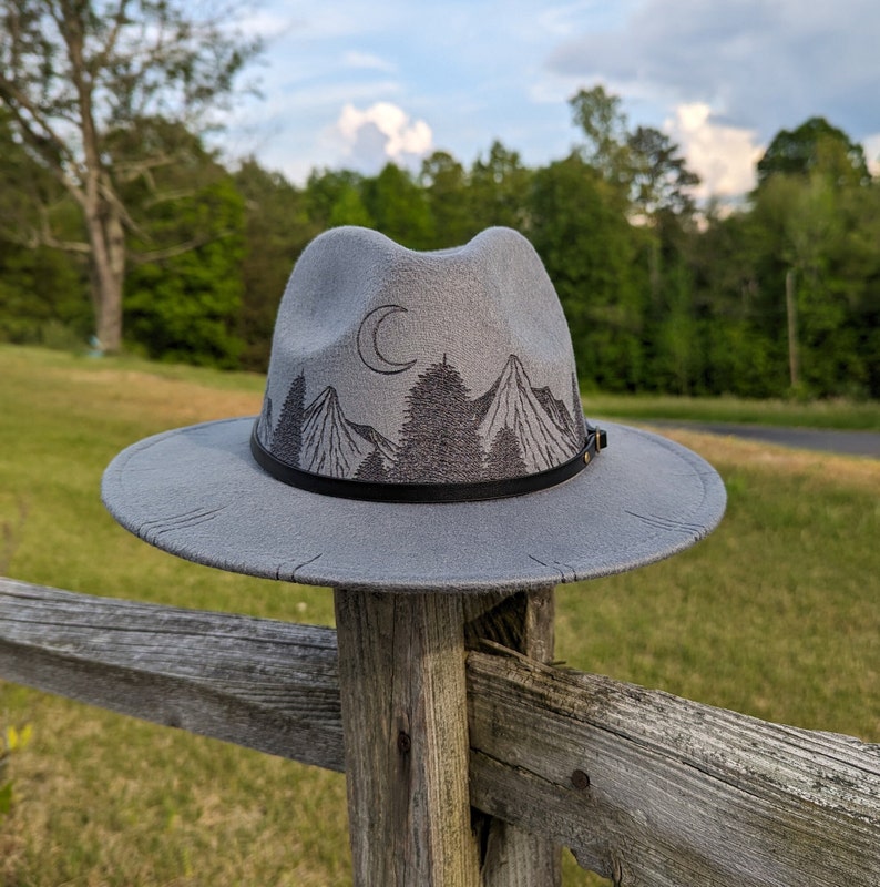 Op de afbeelding: Een grijze wollen fedora hoed met een zwarte band en een geborduurd ontwerp van bergen, bomen en een halve maan.