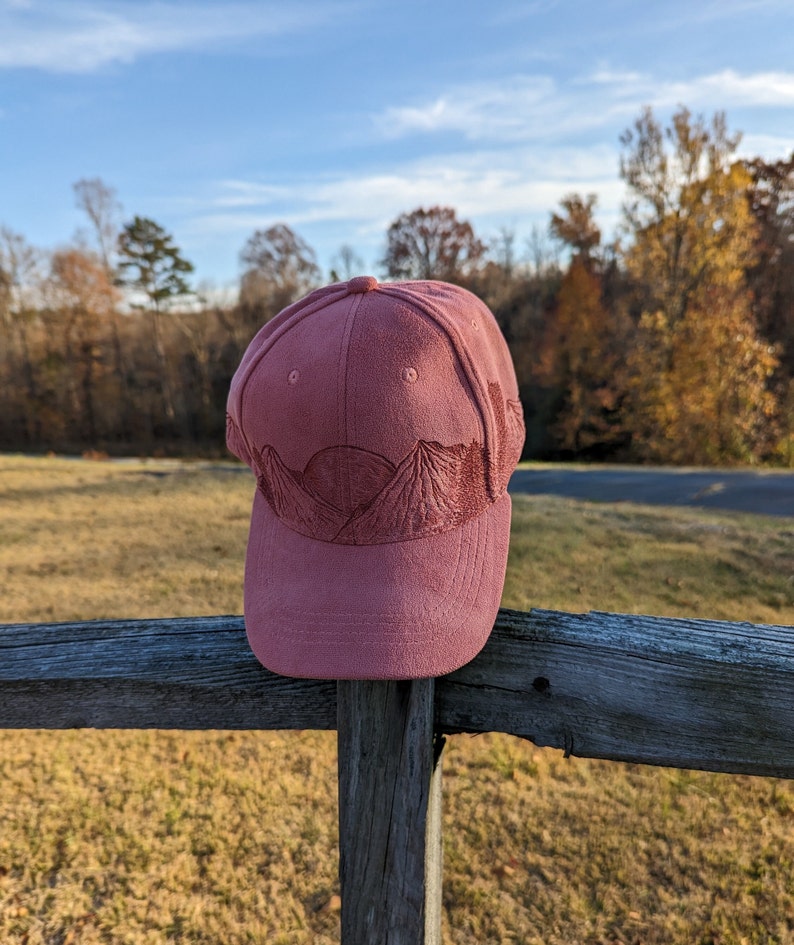 May include: A dusty rose colored baseball cap with a mountain range embroidered on the front. The cap is sitting on a wooden fence.