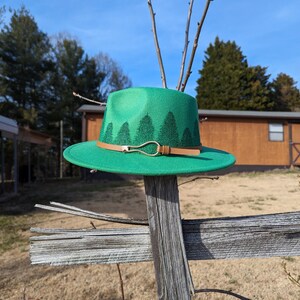 May include: A green felt fedora hat with a brown leather band and a silver buckle. The hat has a design of green trees on the band.