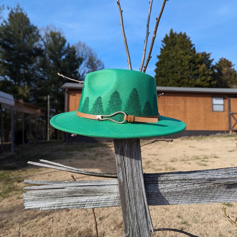 May include: A green felt fedora hat with a brown leather band and a silver buckle. The hat has a design of green trees on the band.