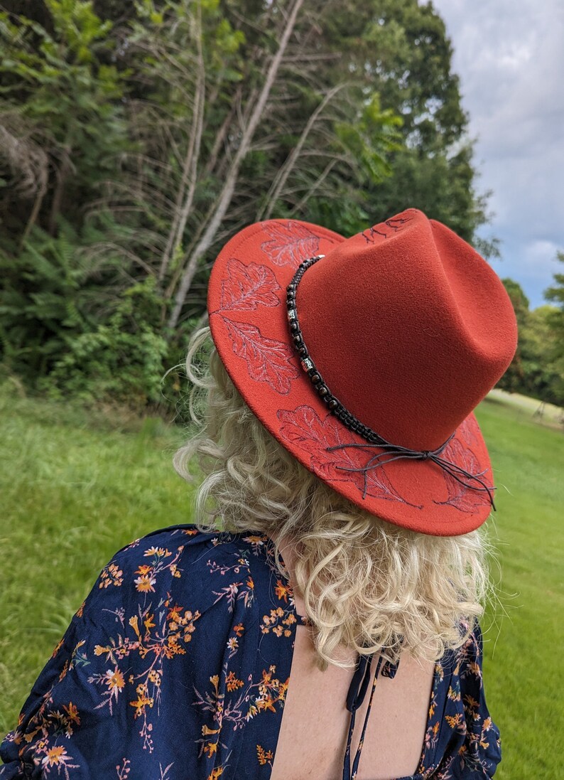May include: A red felt fedora hat with a black leather band and a leaf design. The hat is decorated with a beaded trim.