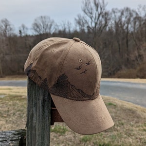 May include: A brown suede baseball cap with a mountain and birds design. The cap has a curved brim and a brown strap.