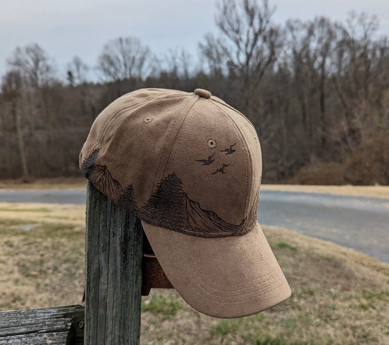 May include: A brown suede baseball cap with a mountain and birds design. The cap has a curved brim and a brown strap.