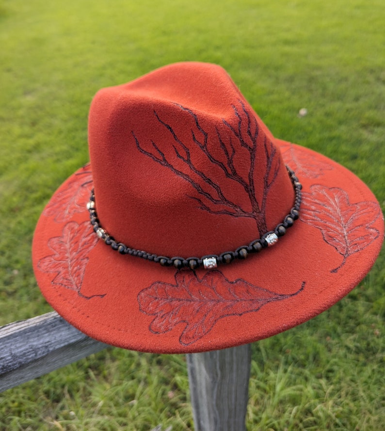 May include: A red felt fedora hat with a black tree design and oak leaf accents. The hat has a brown beaded band with silver accents.