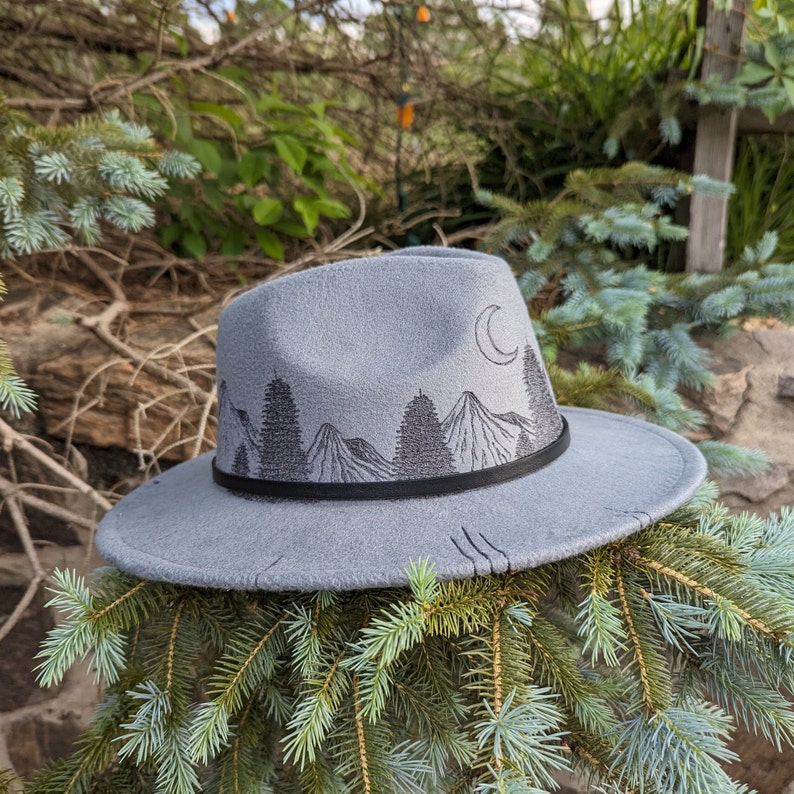 Op de afbeelding: Een grijze wollen fedora hoed met een zwarte band en een geborduurde berg sc&egrave;ne op de voorkant. De sc&egrave;ne bevat een halve maan, bomen en bergen.
