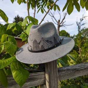 Op de afbeelding: Een grijze wollen fedora hoed met een zwarte band en een geborduurde berg sc&egrave;ne op de kroon. De hoed heeft een brede rand en ligt op een houten hek.