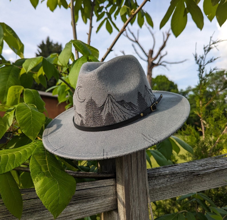Op de afbeelding: Een grijze wollen fedora hoed met een zwarte band en een geborduurde berg sc&egrave;ne op de kroon. De hoed heeft een brede rand en ligt op een houten hek.