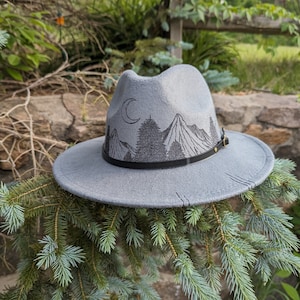 Op de afbeelding: Een grijze wollen fedora hoed met een zwarte leren band en een geborduurd berglandschap aan de voorkant. De sc&egrave;ne bevat een halve maan, bomen en bergen.