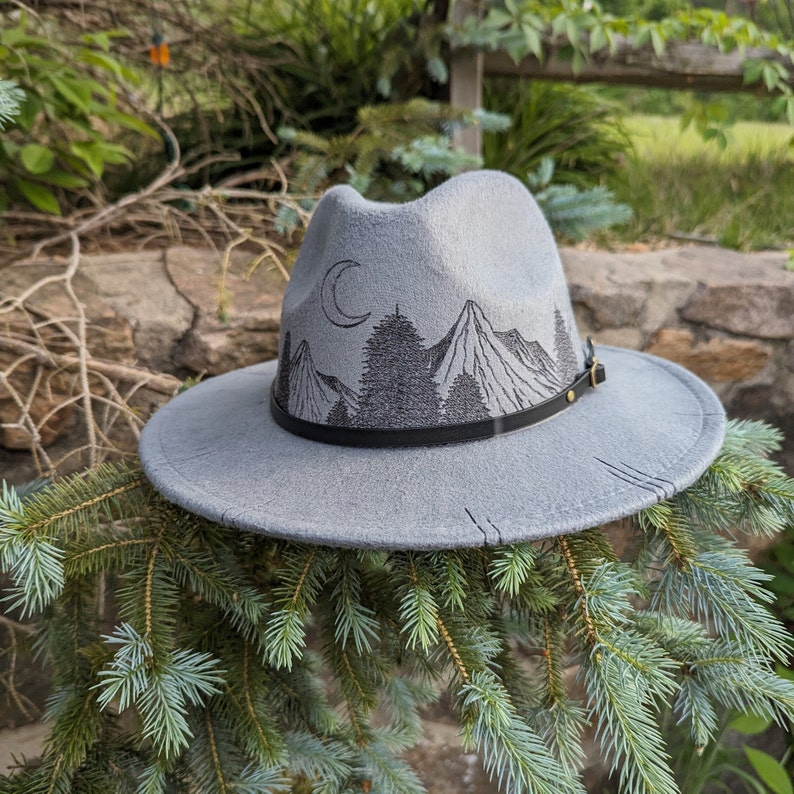 Op de afbeelding: Een grijze wollen fedora hoed met een zwarte leren band en een geborduurd berglandschap aan de voorkant. De sc&egrave;ne bevat een halve maan, bomen en bergen.