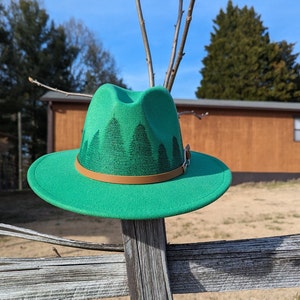 May include: A green felt fedora hat with a brown leather band and a forest scene design. The hat is sitting on a wooden fence.